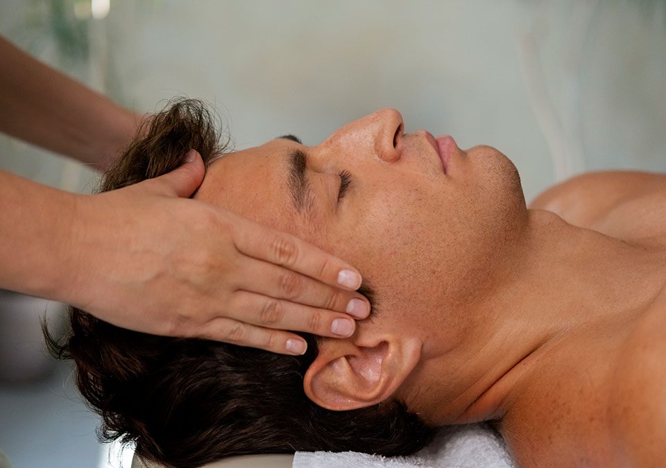 Hombre tumbado en camilla con los ojos cerrados y unas manos a los lados de su cabeza