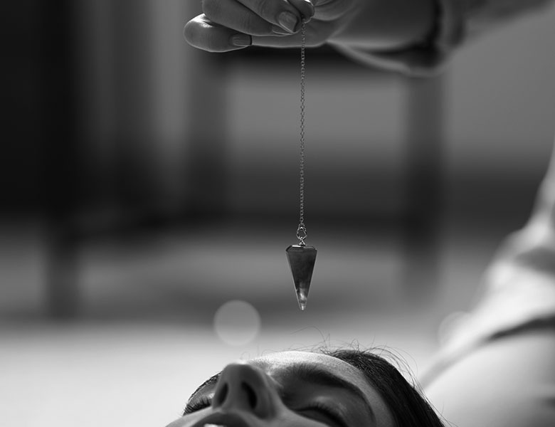 Una mujer tumbada en el suelo y otra persona sostiene una cadena con un mineral en la punta, sobre su frente